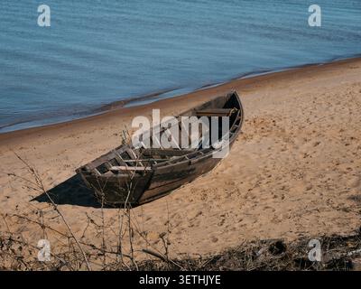 Verwittertes Holzruderboot liegt an einem Sandstrand. Ruhiges blaues Wasser reicht bis zum Horizont. Sanfte Wellen umrunden das Ufer. Die Szene fühlt sich friedlich und qu an Stockfoto