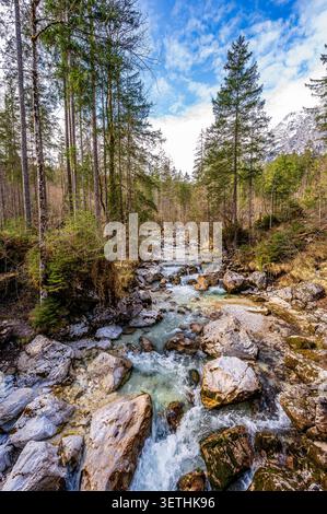 Bergwaldfluss mit klarem türkisfarbenem Wasser, das über Felsen und Felsbrocken in den Berchtesgadener Alpen fließt. Hohe immergrüne Bäume säumen das roc Stockfoto