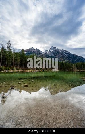 Klarer Alpensee mit sichtbarer seitiger Küste, grünem Wasser und Wald, der schneebedeckte Berggipfel unter einem bewölkten Himmel in der Nähe von Berchtesgaden reflektiert Stockfoto