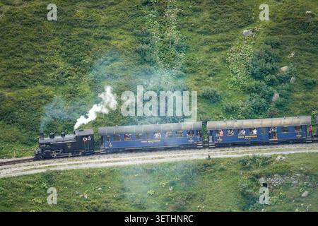Aus der Vogelperspektive der historischen Dampflokomotive der Furka Steam Railway, die blaue Personenwagen durch eine üppig grüne Berglandschaft zieht, Swit Stockfoto