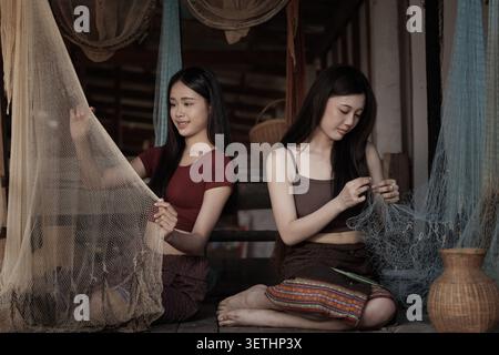 Zwei junge Frauen in traditioneller Kleidung sitzen auf einem Holzboden und reparieren Fischernetze, die kulturelle Handwerkskunst und Kulturerbe in einer ruhigen Umgebung zeigen. Stockfoto