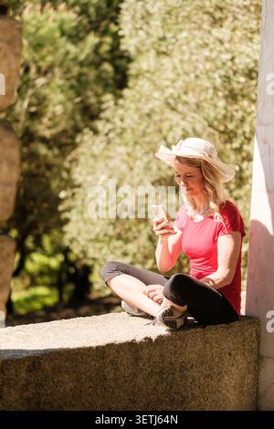 Frau, die auf einer Steinbank sitzt und ihr Smartphone in einem Park benutzt Stockfoto