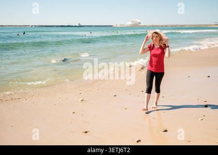 Frau in rotem Hemd und Hut, die an einem sonnigen Tag barfuß an einem Sandstrand entlang läuft Stockfoto