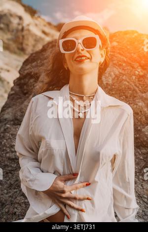 Frau Sommer Strand lächelnde Frau in weißem Hemd und Sonnenbrille posiert vor felsiger Küste Hintergrund bei Sonnenuntergang Stockfoto