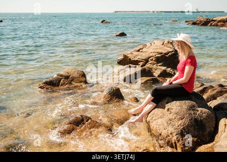 Frau, die auf einem Felsen sitzt und das Meer an einem klaren Sommertag beobachtet Stockfoto