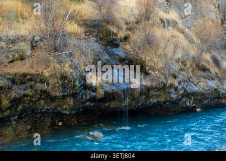 Blick auf einen kleinen Wasserfall, der über ein felsiges, grasbewachsenes Ufer in einen lebhaften blauen Fluss im Khunjerab National Park, Khunjerab, Gilgit, Pakistan stürzt Stockfoto