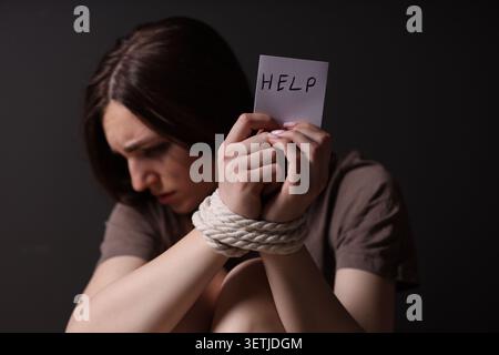 Menschenhandel. Verzweifelte Frau mit gefesselten Händen, die Papier mit Worthilfe auf schwarzem Hintergrund hält Stockfoto