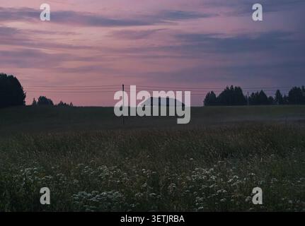 Ländliche Landschaft mit blühenden Wildblumen, einem Bauernhaus und einem farbenfrohen Abendhimmel bei Sonnenuntergang im Sommer Stockfoto