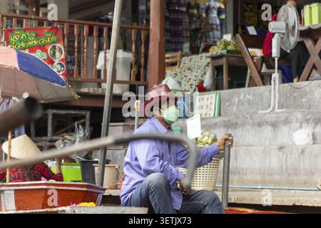 Bangkok, Bangkok, Thailand - 21. Juli 2026: Blick auf einen Mann mit Cowboyhut und Gesichtsmaske, der in einem Holzboot auf einem schwimmenden Markt sitzt. Stockfoto