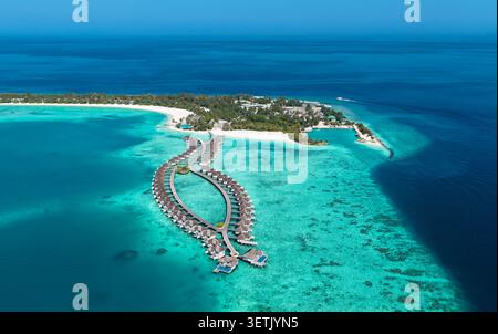 Blick aus der Vogelperspektive auf die Bungalows über dem Wasser, die in einzigartiger Form über einer türkisfarbenen Lagune mit Korallenriffen und einer tropischen Insel mit weißen Sandstränden angeordnet sind Stockfoto