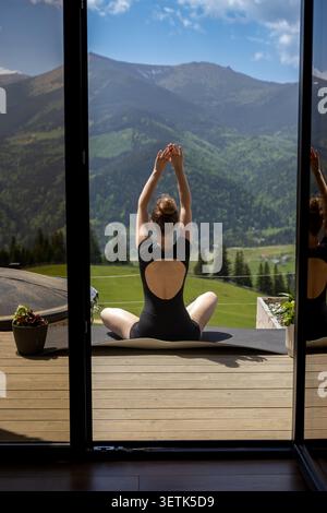 Frau, die sich auf der Terrasse in den Karpaten ausdehnt. Frau sitzt auf einer Yogamatte mit erhobenen Armen auf einer Holzterrasse mit Blick auf die grüne Ukrainisch Stockfoto