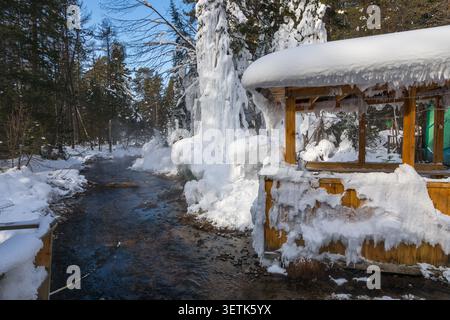 Blick auf die heiße Quelle Goryachiye Istochniki Khakusy, die durch einen verschneiten Wald fließt, mit einem hölzernen Unterschlupf in Eis im Severobaykal'skiy Rayon, Respu Stockfoto