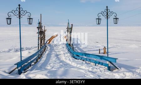 Blick auf Goryachiye Istochniki Khakusy mit einem schneebedeckten Pier mit blauen Geländern, der in den gefrorenen Baikalsee im Severobaykal'skiy Rayon, R, reicht Stockfoto