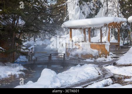 Blick auf Goryachiye Istochniki Khakusy mit einer Holzbrücke über eine dampfende Thermalquelle, schneebedeckten Bäumen und einem hölzernen Pavillon in Severobaykal'Ski Stockfoto