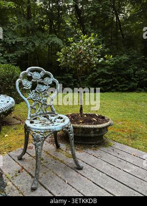 Vintage schmiedeeiserner Gartenstuhl mit Rosenmuster auf einer hölzernen Terrasse neben einem Topfbaum Stockfoto