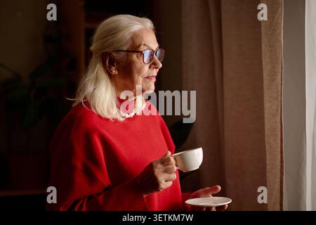 Seniorin hält Kaffeetasse und schaut zu Hause durch das Fenster Stockfoto
