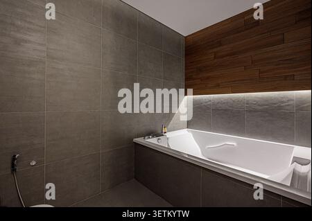Das moderne Bad verfügt über graue geflieste Wände, eine weiße Badewanne und eine holzgetäfelte Wand mit Beleuchtung Stockfoto