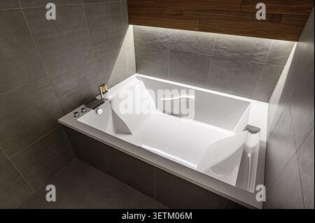 Das moderne Bad verfügt über eine elegante, weiße Badewanne mit dunkelgrauen Fliesen. Holzakzente und dezente Beleuchtung schaffen ein Spa-ähnliches Ambiente Stockfoto