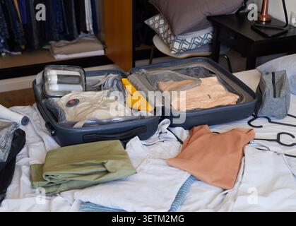 Ein offener Koffer auf einem ungebauten Bett ist organisiert mit Staubsaugertaschen, gefalteter Kleidung, Toilettenartikeln, einer gelben Tasche und zusätzlichen Kleidungsstücken, die für trave bereit sind Stockfoto