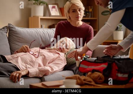 Seniorin ruht auf Sofa, während die Betreuerin den Hausbesuch vorbereitet Stockfoto