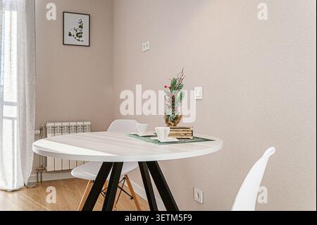Gemütliche Einrichtung mit einem weißen runden Tisch für zwei Personen, geschmückt mit einem kleinen Weihnachtsbaum. Helle, neutrale Wände und minimalistische Einrichtung schaffen eine ruhige Atmosphäre Stockfoto