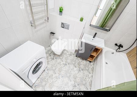 Modernes Badezimmer mit weißer Waschmaschine, Toilette, Waschbecken mit Schrank und Badewanne. Mit hellgrauen Fliesen und gemustertem Boden Stockfoto