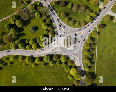 Aus der Vogelperspektive direkt über einem von Bäumen gesäumten Verkehrskreis oder Kreisverkehr mit Autos und Fahrzeugen in Bewegung zur Hauptverkehrszeit durch die Landschaft mit grünem Gras Stockfoto