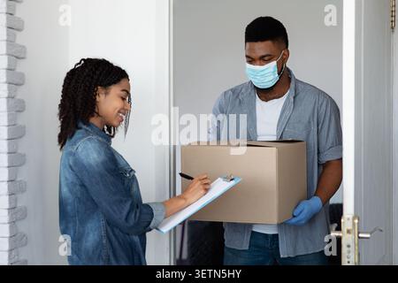 Sichere Lieferung Nach Hause. Schwarze Frau Erhielt Parcel Von Courier In Medizinischer Maske Stockfoto