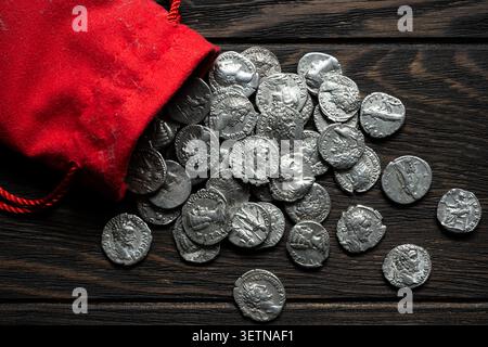 Ein Blick von oben auf eine hellrote Tüte mit Kordelzug aus Stoff, die einen Stapel alter Silbermünzen auf einen dunklen, rustikalen Holztisch verschüttet. Die Münzen sind mit Detai versehen Stockfoto