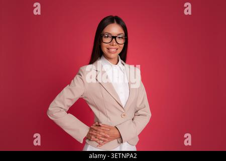 Selbstbewusste Geschäftsfrau mit Brille und Blazer vor rotem Hintergrund mit Kopierraum, professionellem Porträt, Führungs- und Karriereerfolg Stockfoto