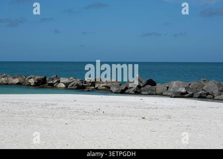 Sandstrand mit grobem weißem Sand führt zu einem felsigen Wellenbrecher, der sich bis in den Golf erstreckt. Stockfoto