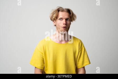 Junger blonder Mann in einem gelben T-Shirt, der Schock und Überraschung ausdrückt, Augen und Mund auf hellgrauem Hintergrund weitet und unerwartete Nachrichten erkennt Stockfoto