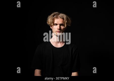 Junger Mann mit blonden Haaren und ernstem Ausdruck posiert im Studio mit dramatischer Beleuchtung und schwarzem Hintergrund, symbolisiert Kontemplation, Tiefe und Stockfoto