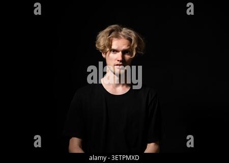 Junger Mann mit hellen Stoppeln und blonden lockigen Haaren, mit einem schwarzen T-Shirt, stehend vor dunklem Hintergrund, mit verwirrtem und intensivem Blick Stockfoto