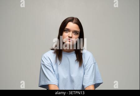 Junge Frau mit braunen Haaren, die blaues T-Shirt trägt, die Kamera in einem Studio ansieht, grauer Hintergrund Stockfoto