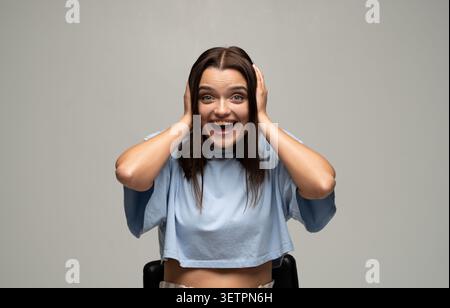 Junge Frau, die die Ohren mit den Händen bedeckt, mit offenem Mund Überraschung und Freude ausdrückt, staunend und aufgeregt Stockfoto