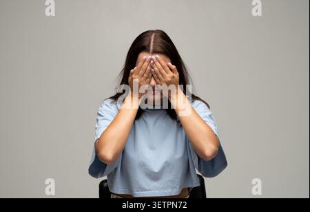 Junge Frau, die das Gesicht mit den Händen bedeckt, sich ängstlich fühlt, isoliert ist und sich schämt auf grauem Hintergrund Stockfoto