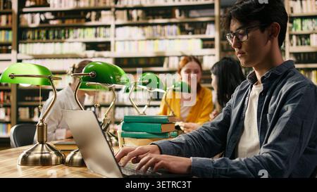 Junger Mann, der auf einem Laptop in der Bibliothek schreibt, studiert und online lernt, Klassenkameraden arbeiten in der Nähe mit Büchern und Regalen in weichem Fokus um den Tisch herum. Asiatischer Schüler zeigt digitales Lernen. Stockfoto