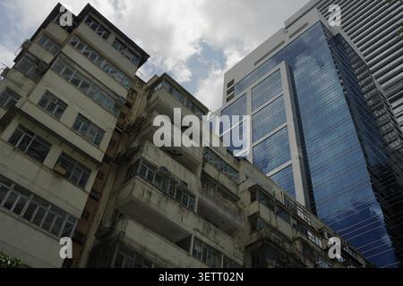 Flacher Blickwinkel, kontrastierende verwitterte Wohnhäuser mit einem eleganten modernen blauen Glashochhaus in Hongkongs dichter Stadtlandschaft. Stockfoto