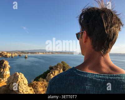 Nahaufnahme eines stilvollen Mannes mit Sonnenbrille, während er die atemberaubende Küstenlandschaft bewundert. Blick über den endlosen Atlantik. Stockfoto