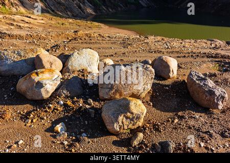 Große Felsbrocken auf sandigem Boden in einem Steinbruch mit einem kleinen See im Hintergrund unter hellem natürlichem Sonnenlicht, die strukturierten Schatten erzeugen Stockfoto