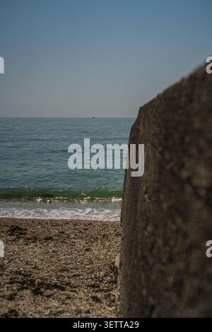 Ein Blick auf die Kieselstrandlinie, die auf das grün getönte Meerwasser und den Horizont dahinter trifft, von einer großen strukturierten Betonmauer, Charl, aus gesehen Stockfoto