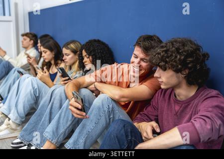 Verschiedene Gruppen junger Studenten sitzen hintereinander, die sich individuell auf ihre Mobiltelefone konzentrieren, während ein lächelnder Mann mit einem anderen interagiert und hebt hervor Stockfoto