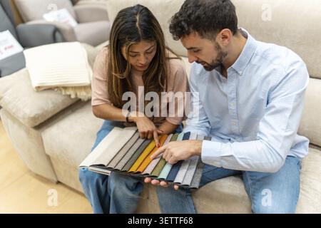 Junges Paar auf einem Showroom-Sofa, das große Stoffmuster und Farbmuster miteinander vergleicht, Polster wählt und Innenarchitektur und Improvisation plant Stockfoto