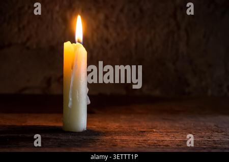 Eine brennende Kerze auf einem rauen Holzregal. Eine zündete Kerze in der Dämmerung. Stockfoto