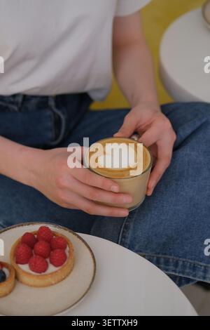Hände halten Latte mit Herzschaum neben Himbeertorte in gemütlicher Café-Umgebung Stockfoto
