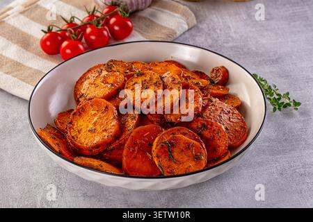 In Scheiben geröstete Süßkartoffeln mit Kräutern in einer weißen Schüssel. Hintergrund ist Knoblauch aus Kirschtomaten auf gestreiftem Tuch. Warme Orangetöne, sanfte Beleuchtung, Stockfoto