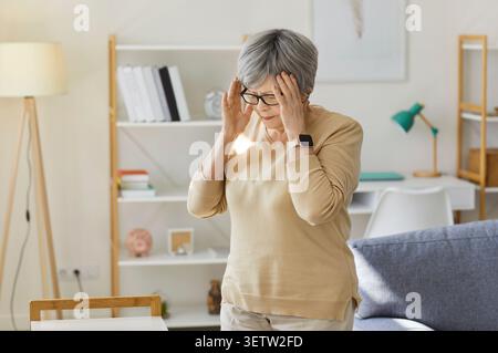 Gestresste ältere Frau mit Brille, die zu Hause im Wohnzimmer steht und an Kopfschmerzen leidet Stockfoto
