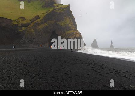Dramatischer schwarzer Sandstrand von Reynisfjara in Island mit Basaltsäulen, riesigen Meeresstapeln, Meereswellen und Touristen unter einem bewölkten Himmel. Stockfoto