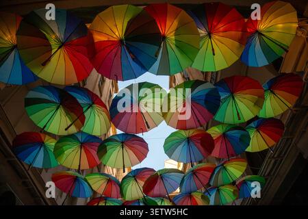 Bunte Regenschirme hängen über einer belebten Straße und werfen verspielte Schatten, während die Menschen an einem klaren, sonnigen Tag unter ihnen spazieren gehen. Stockfoto
